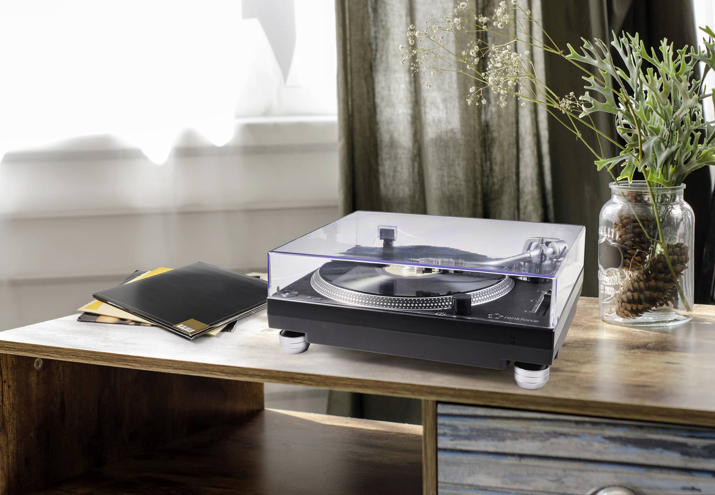 Tourne-disque posé sur une table à côté d'une pile de disques vinyles. À l'arrière-plan, des vases en verre contenant des plantes sont disposés.