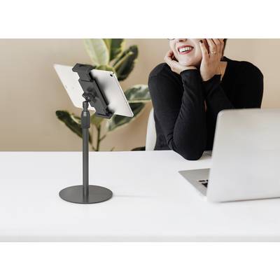 Une personne est assise à une table et sourit tout en regardant un ordinateur portable et une tablette fixée sur un support. Une plante se trouve en arrière-plan.