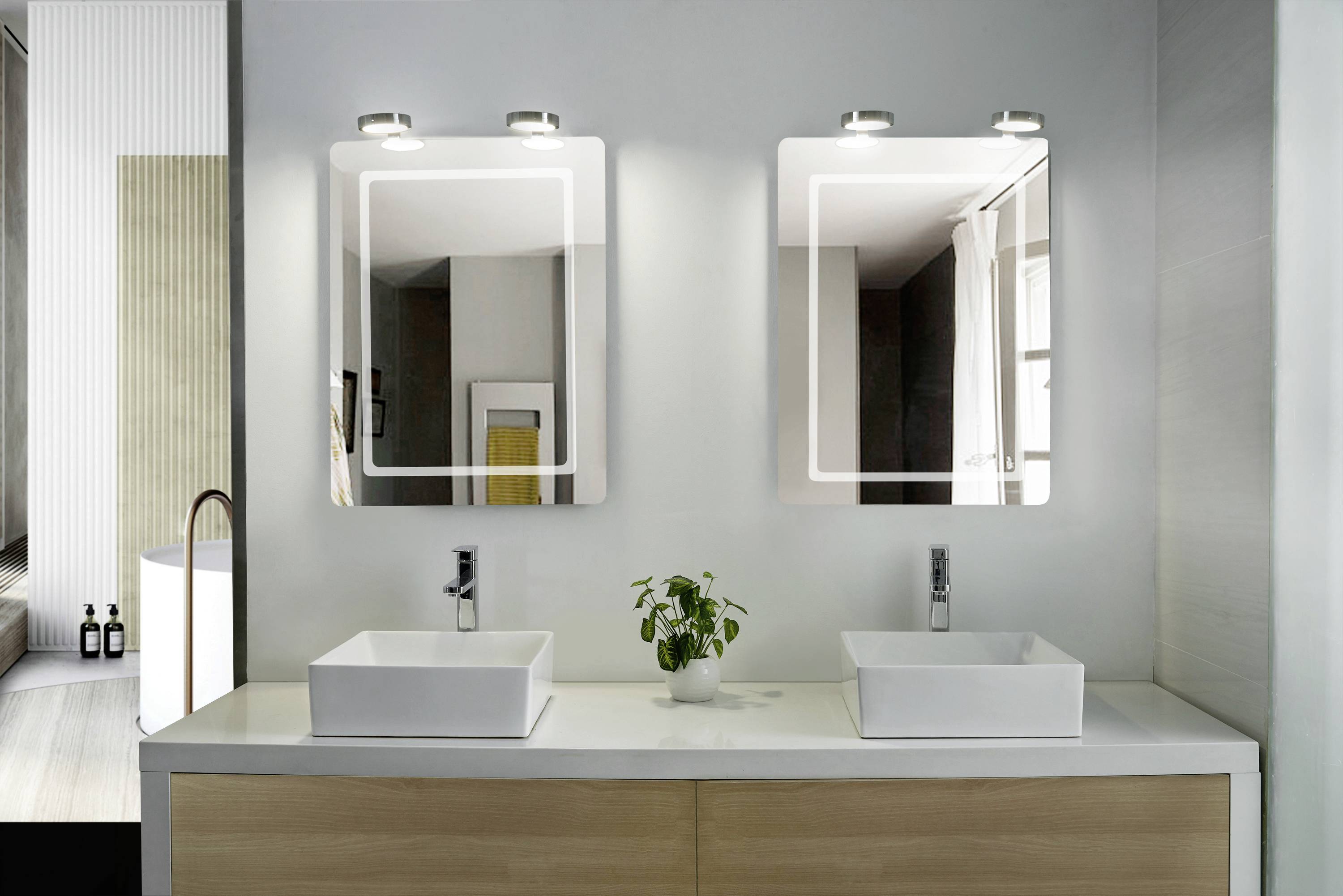 Une salle de bain moderne avec deux miroirs éclairés, deux lavabos blancs et une plante entre eux. Une baignoire est visible en arrière-plan.