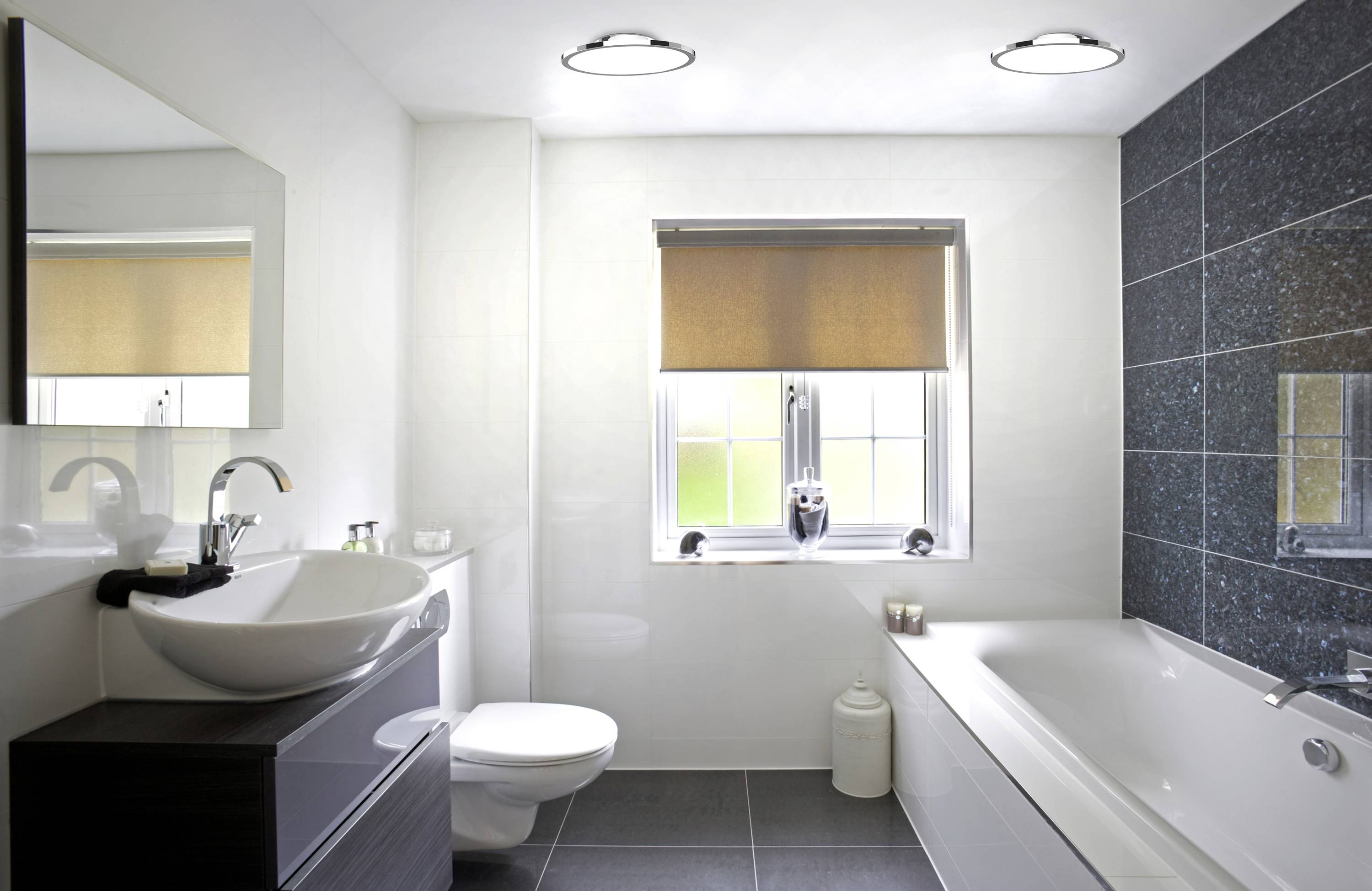 Salle de bain moderne avec lavabo blanc, carrelage gris foncé, baignoire et fenêtre avec stores. Éclairage lumineux.