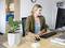 Une femme est assise souriante à son bureau et travaille sur un ordinateur. À côté d'elle se trouve une plante et un verre d'eau. Des étagères sont visibles en arrière-plan.