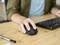 Une personne utilise une souris d'ordinateur sur une table en bois, à côté d'un clavier et d'un stylo. La scène représente un travail de bureau.