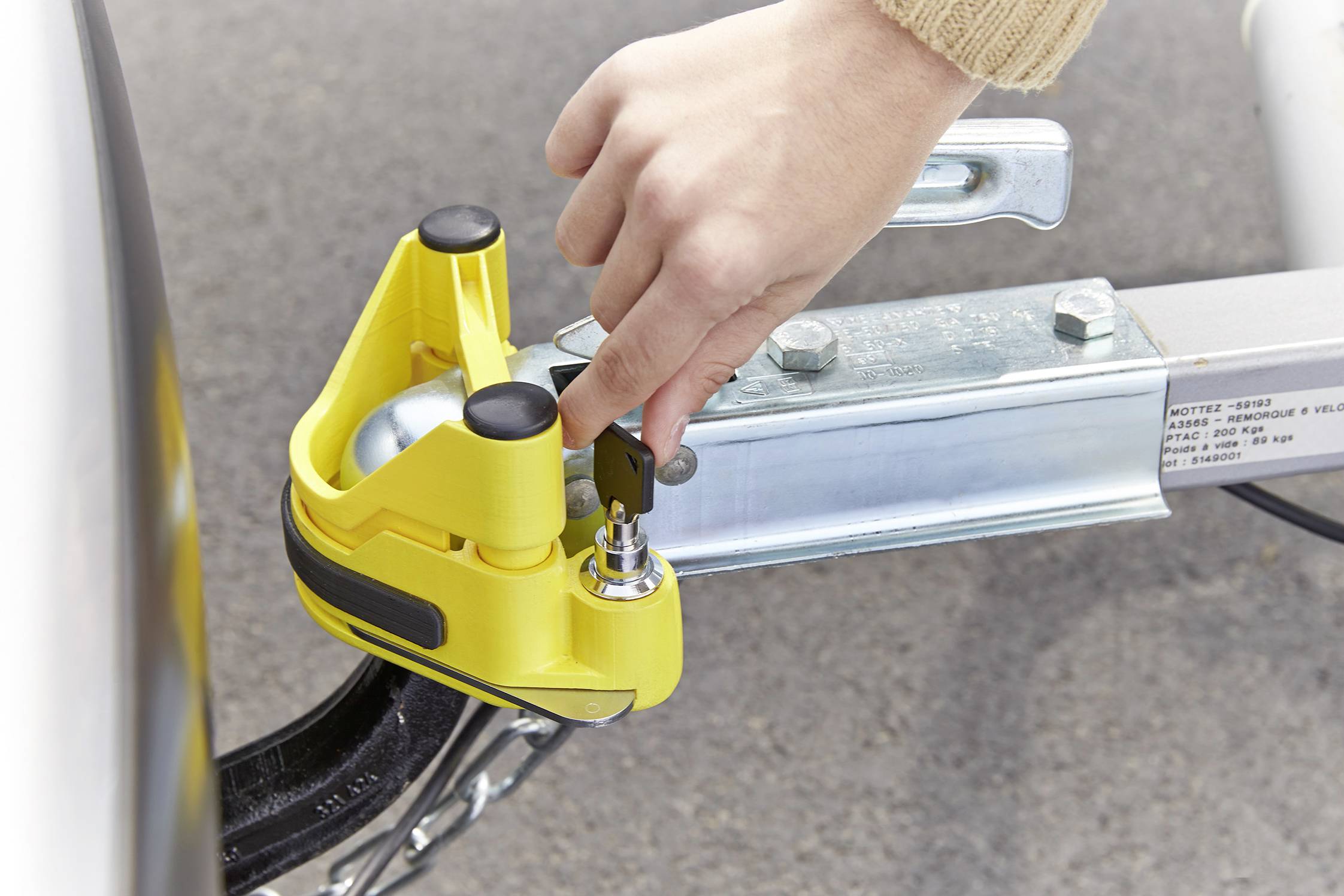 'Une main verrouille un cadenas jaune sur un attelage de remorque. Le cadenas de sécurité protège contre le vol.'