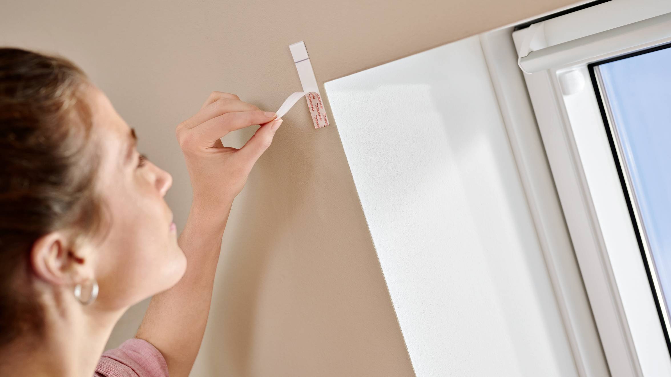 Une femme accroche un crochet blanc sur un mur beige près d'une fenêtre, probablement pour y suspendre des objets.
