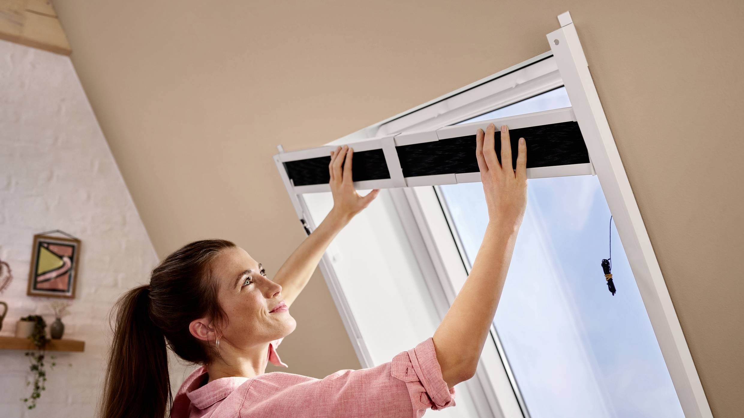 Une femme installe un moustiquaire sur une fenêtre ouverte. Elle regarde avec concentration et travaille soigneusement pour le fixer.