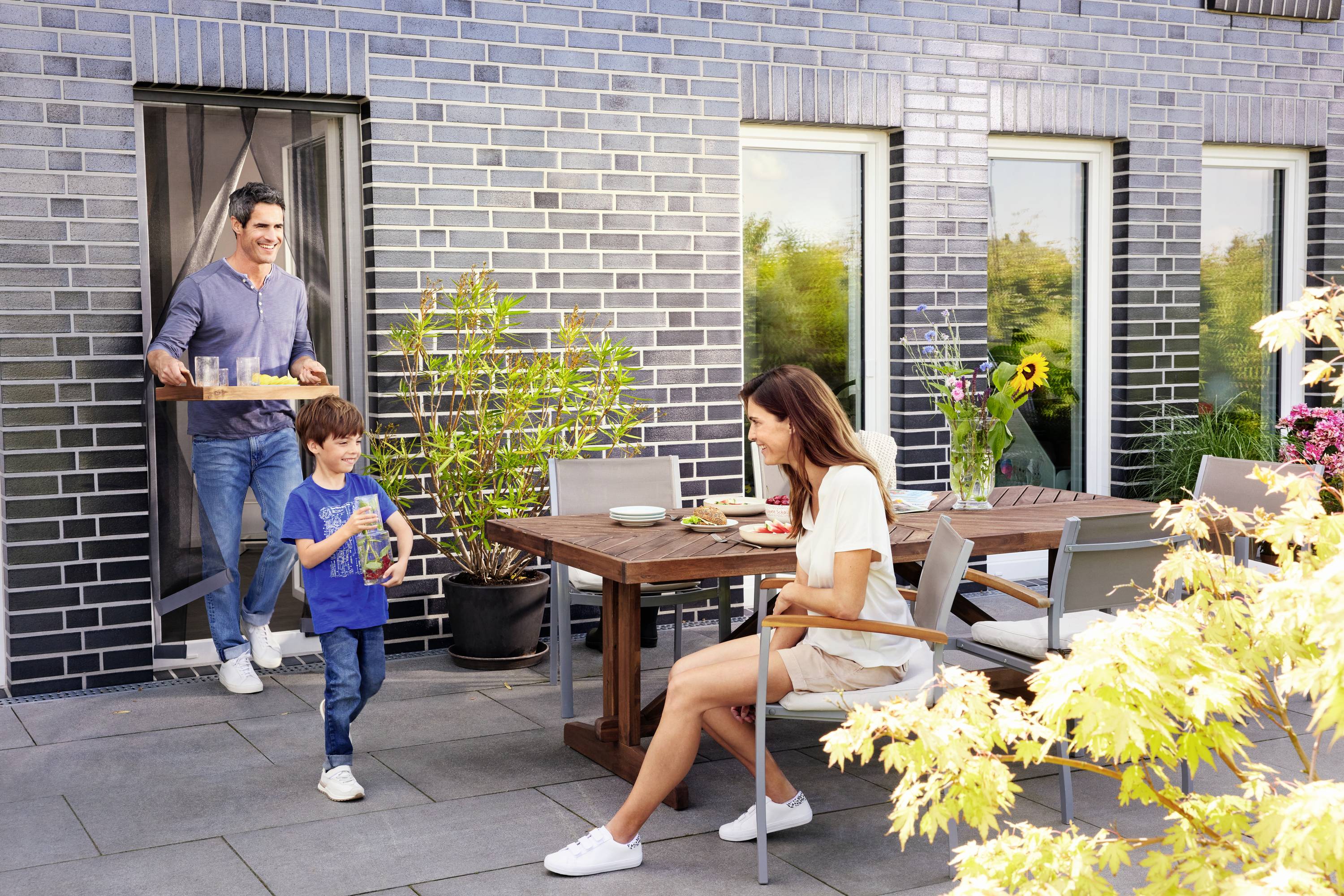 Une famille mange sur une terrasse. Le père apporte un plateau, le fils tient un verre, et la mère est assise à table.
