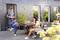 Une famille mange sur une terrasse. Le père apporte un plateau, le fils tient un verre, et la mère est assise à table.