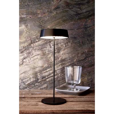 Une élégante lampe de table noire sur une table en bois, à côté d'un verre vide posé sur une assiette carrée, devant un mur en pierre.