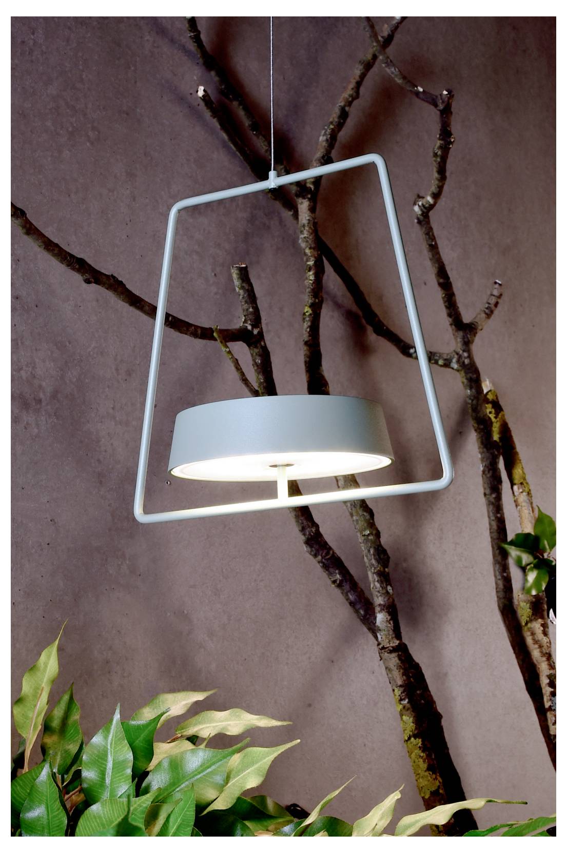 Suspension moderne au cadre géométrique blanc et à l'abat-jour bleu pâle, présentée devant un mur d'accent orné de branches aux feuilles vertes en partie basse.