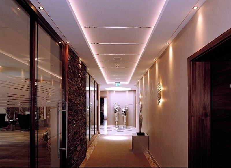 Couloir moderne, bien éclairé, avec des murs décoratifs et des arrangements floraux. Une porte en bois à droite, une paroi vitrée à motifs rayés à gauche.