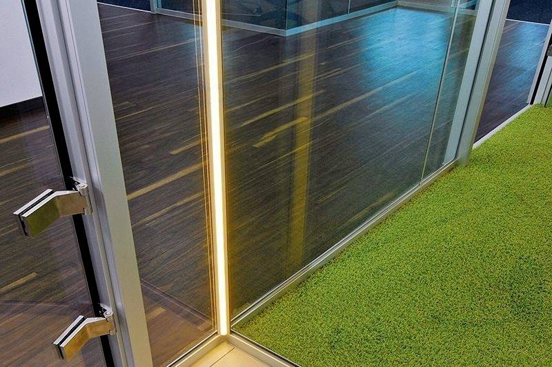Cloison vitrée dans un bureau moderne avec un sol en gazon artificiel vert et une bande lumineuse discrète sur le mur.