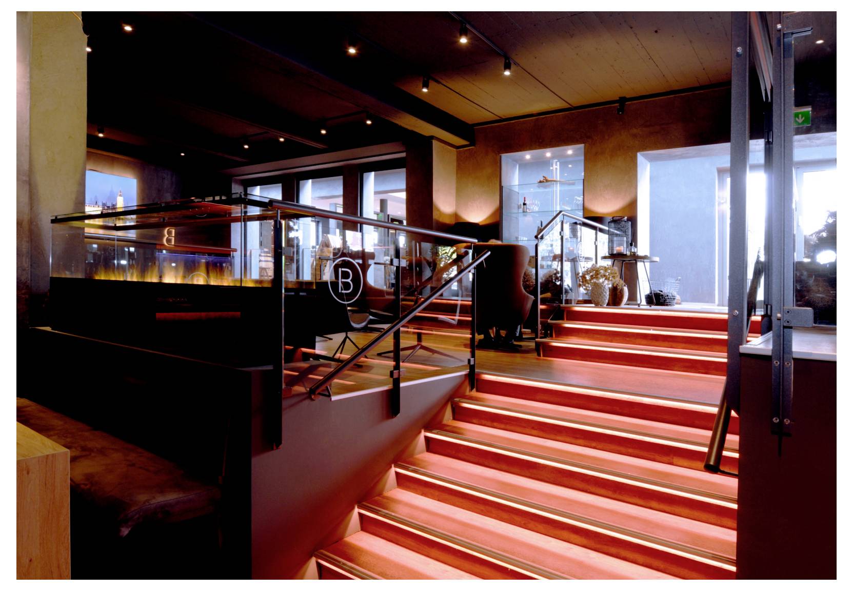 Intérieur moderne avec un escalier éclairé de lumière rouge menant à un bar ou à un salon avec des garde-corps en verre et des assises.