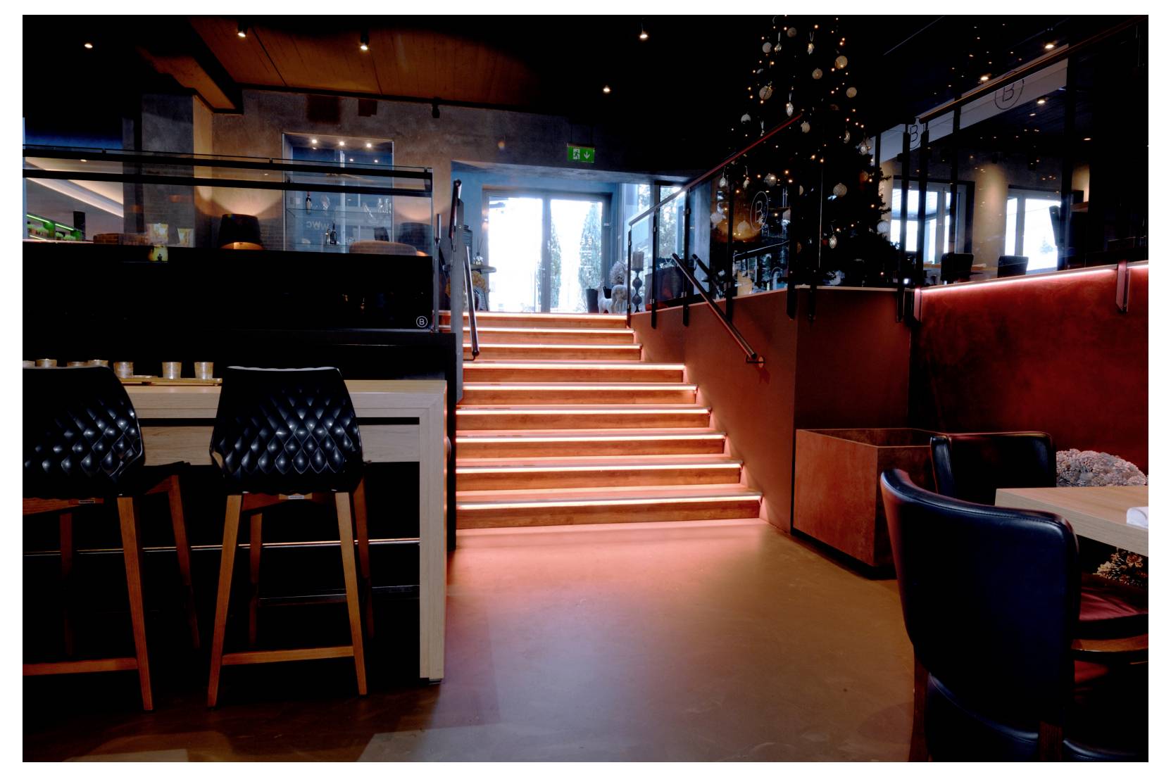 Intérieur de restaurant élégant avec un escalier lumineux menant à une entrée éclatante, encadré de tabourets de bar, de tables et d'un éclairage d'ambiance chaleureux.