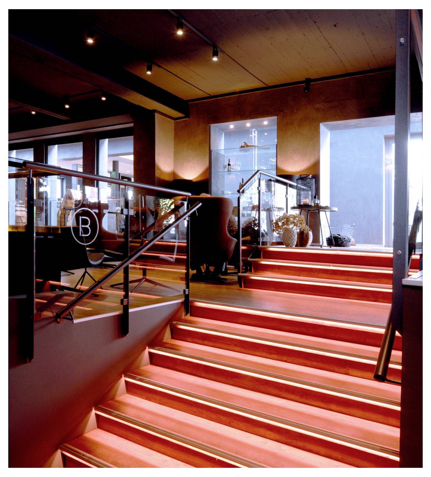 Escalier intérieur moderne aux marches éclairées en rouge, avec garde-corps en verre et une porte lumineuse en haut, dans un hall ou un hôtel élégant.