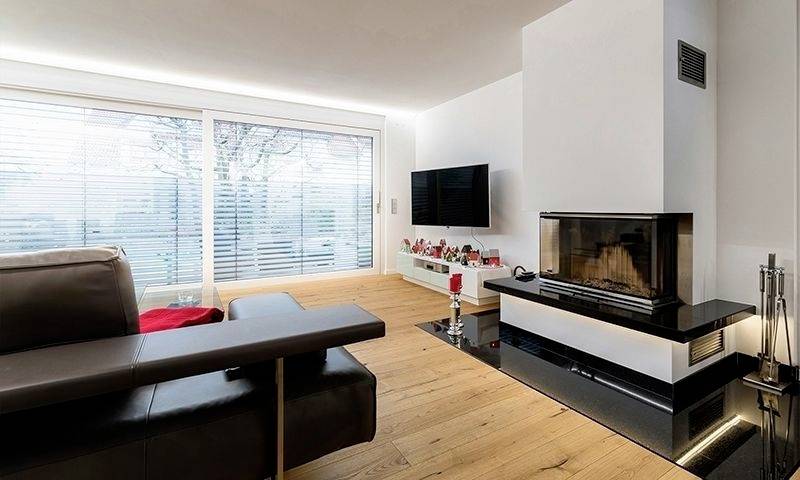 Salon moderne avec cheminée, canapé en cuir, téléviseur à écran plat fixé au mur, sol en bois et grandes fenêtres équipées de stores.
