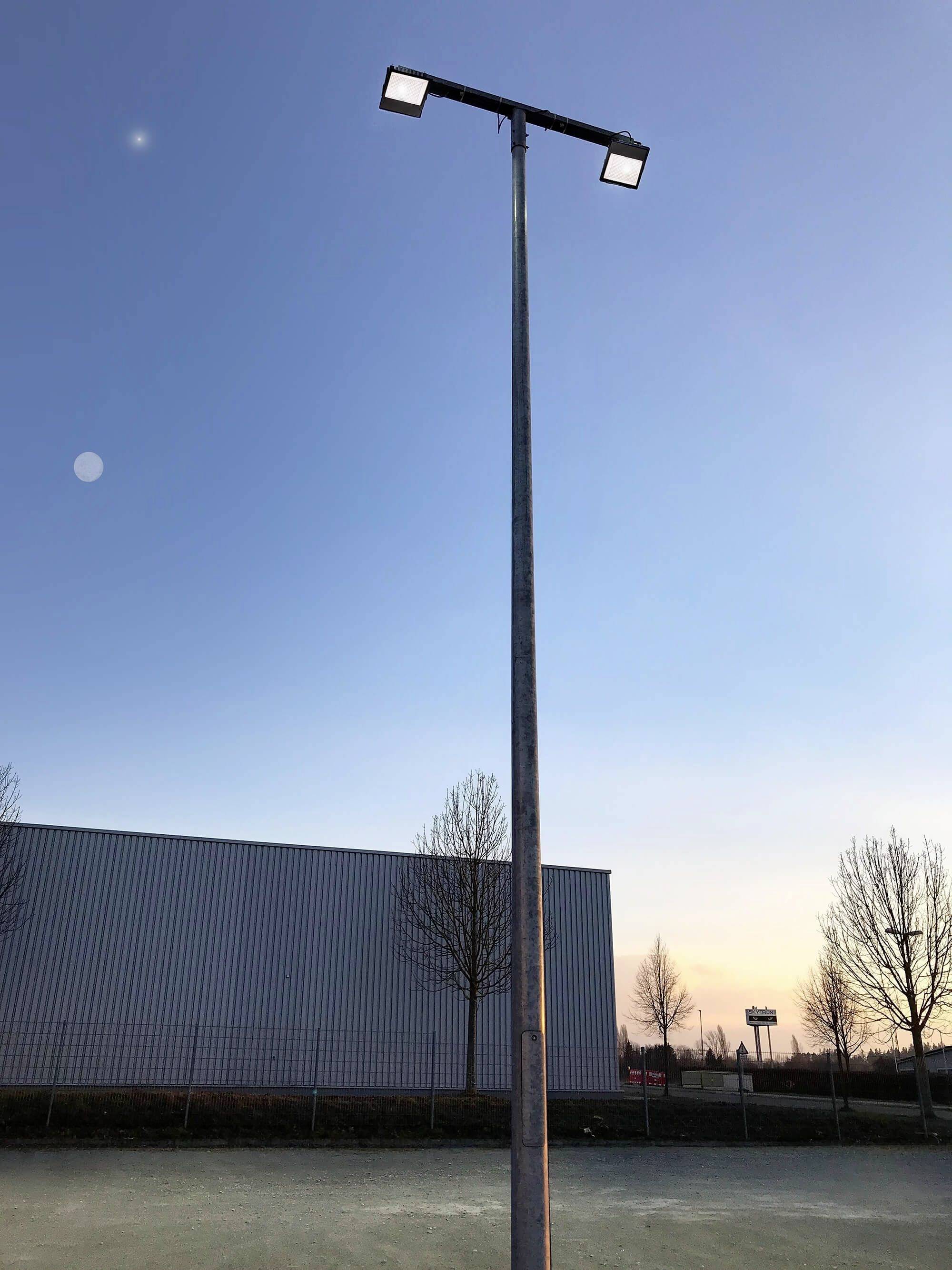 Un lampadaire élevé se dresse sur une place à côté d'un bâtiment moderne. Le ciel, au crépuscule, est clair avec quelques nuages.