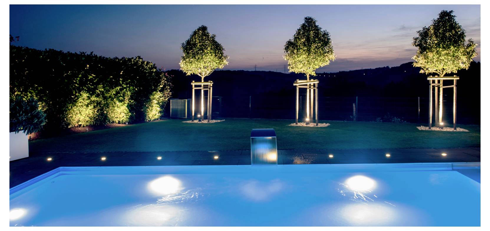 Piscine de jardin illuminée au crépuscule, avec un éclairage paysager sur les arbres et la pelouse, créant une scène extérieure paisible et nocturne.