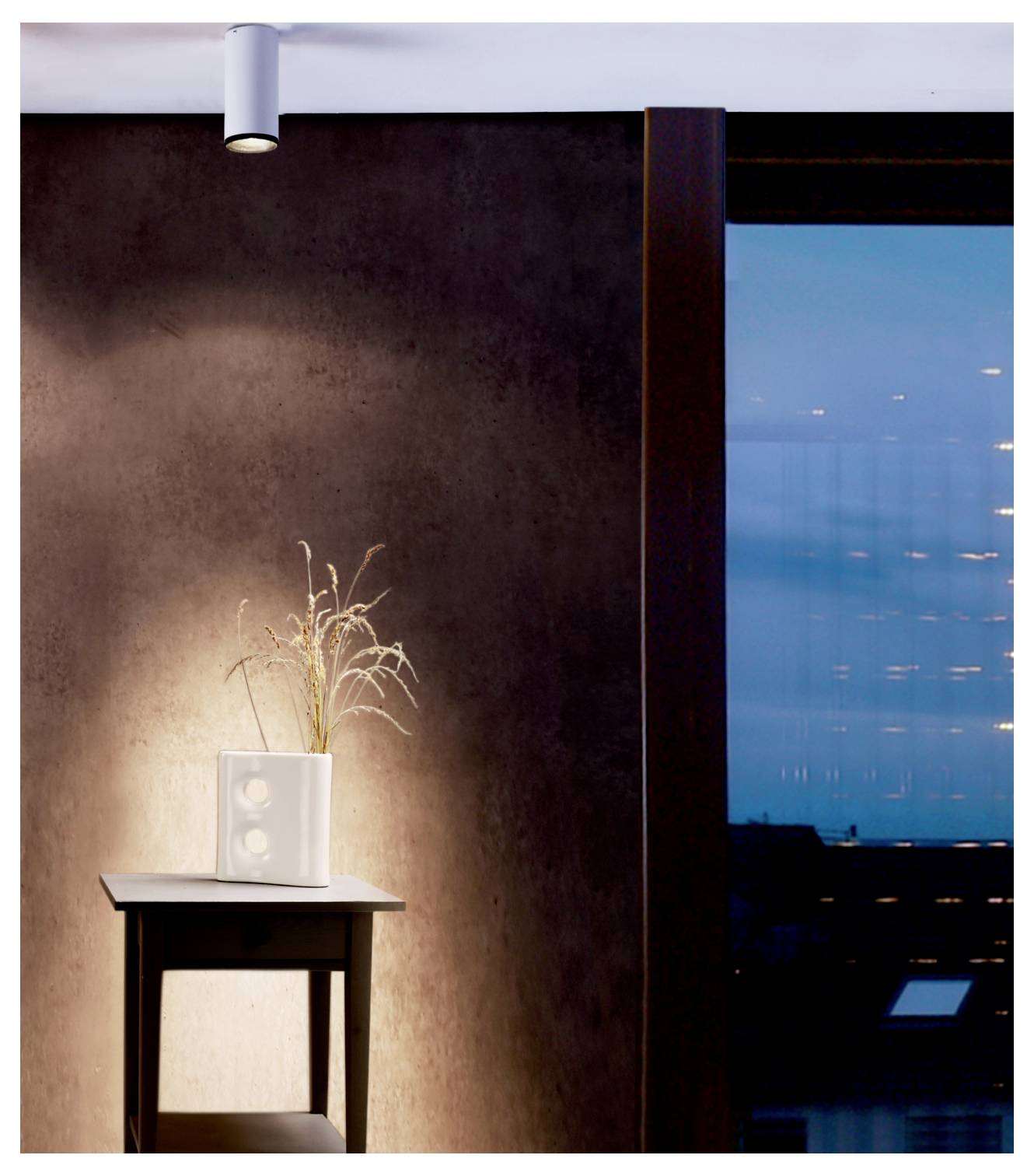 Intérieur épuré avec une petite table supportant un vase blanc de herbes séchées, éclairé par un spot mural à côté d'une grande fenêtre bleue au crépuscule.