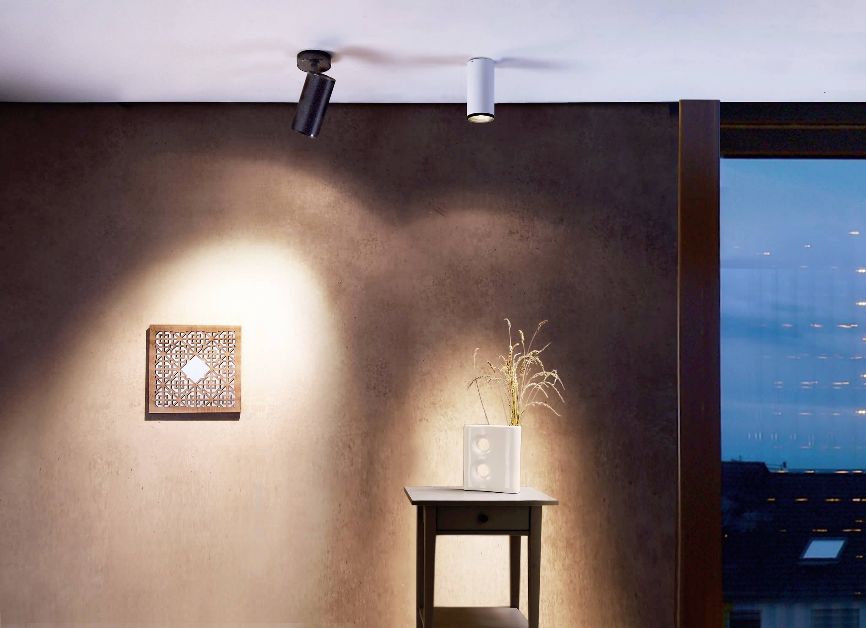 Mur avec éclairage artificiel : Deux luminaires suspendus illuminent un carré artistique et une table avec un vase en verre sur un mur rustique brun.