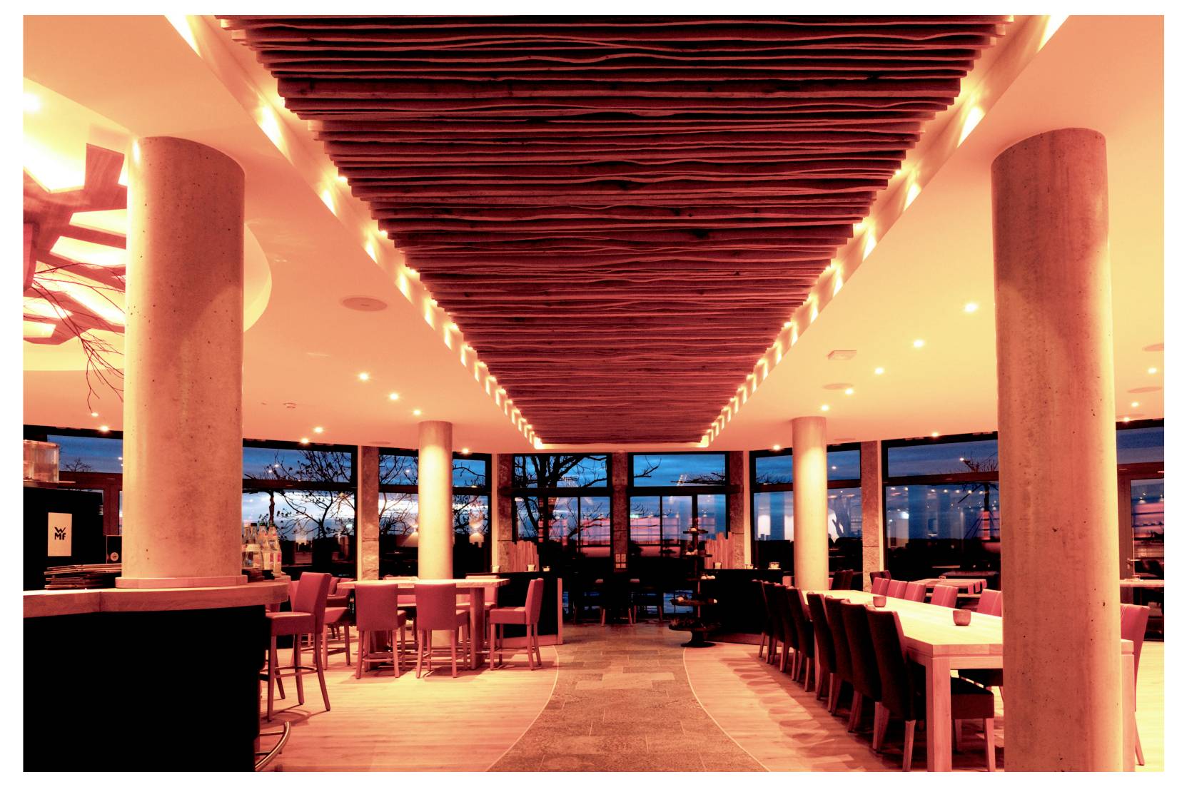 Intérieur de restaurant moderne avec des lattes rouges au plafond, une allée centrale, des tables et des chaises, et de grandes fenêtres donnant sur l'extérieur au crépuscule.