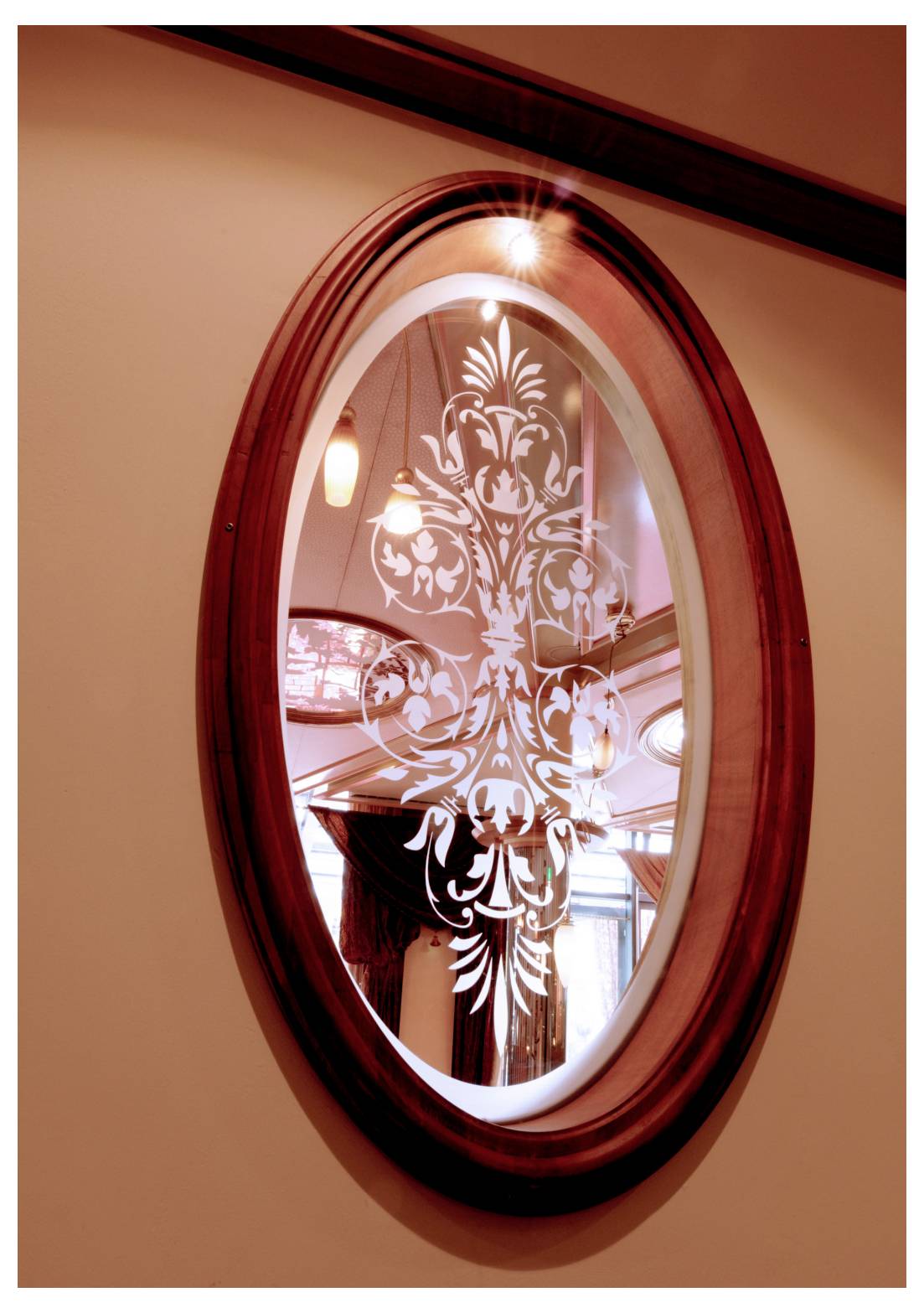 Miroir ovale mural au cadre en bois foncé et au motif floral blanc gravé, reflétant les lumières de la pièce et une fenêtre.