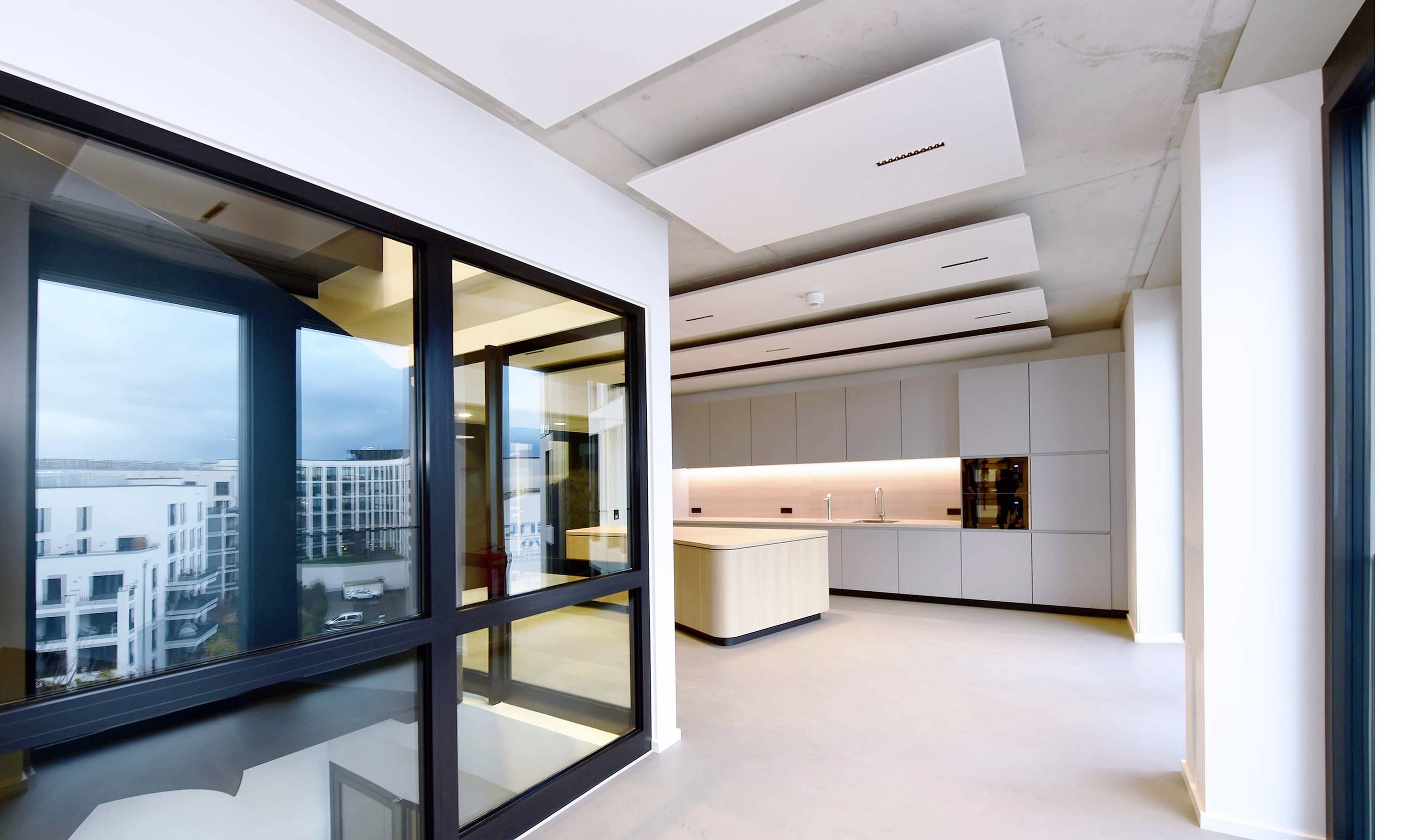 Cuisine moderne et lumineuse avec de grandes fenêtres, four encastré et îlot central dans un espace spacieux offrant une vue sur les immeubles de la ville.