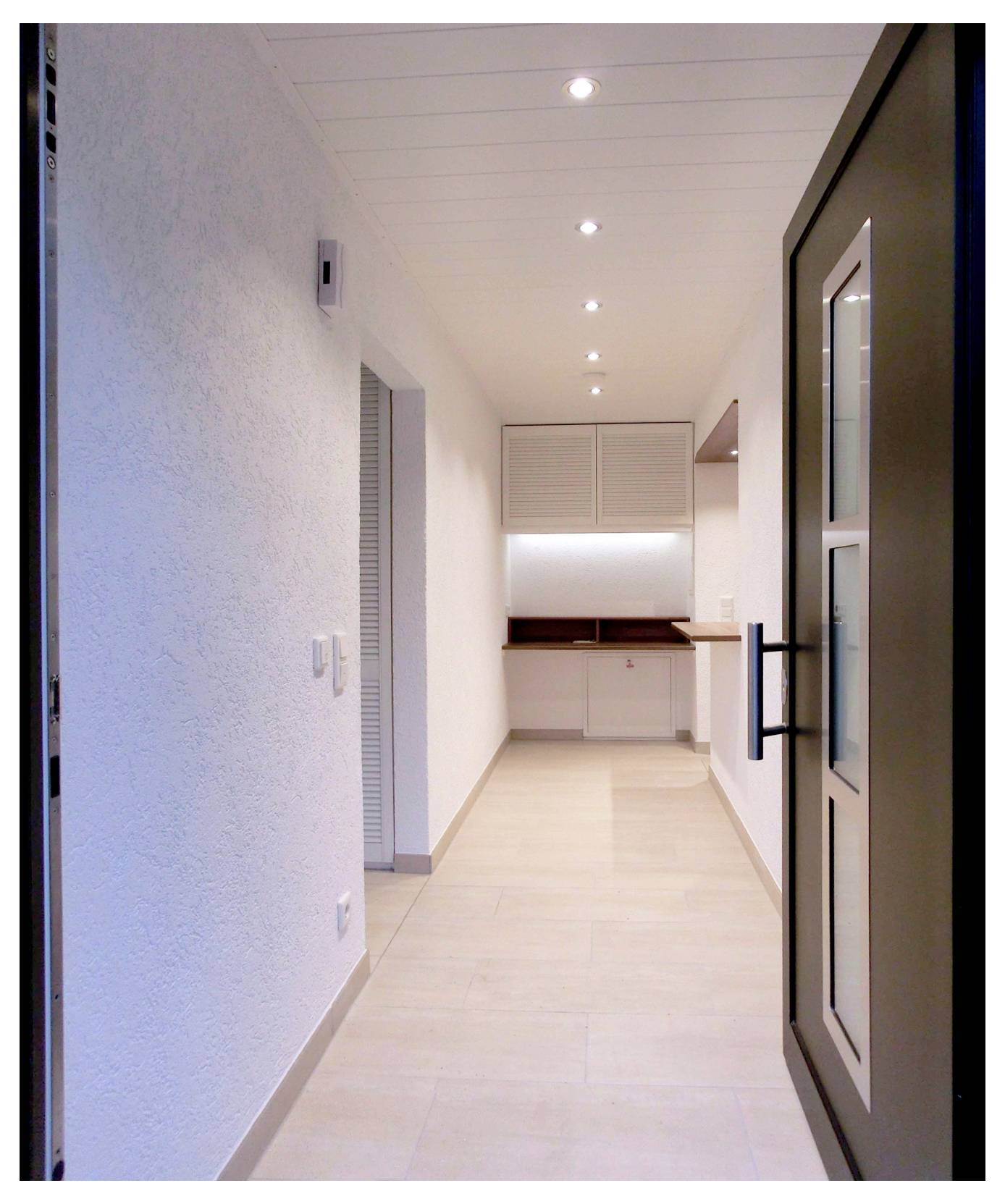 Couloir étroit et lumineux menant à une petite cuisine moderne aux placards blancs et aux spots encastrés au plafond, vu depuis une porte ouverte.