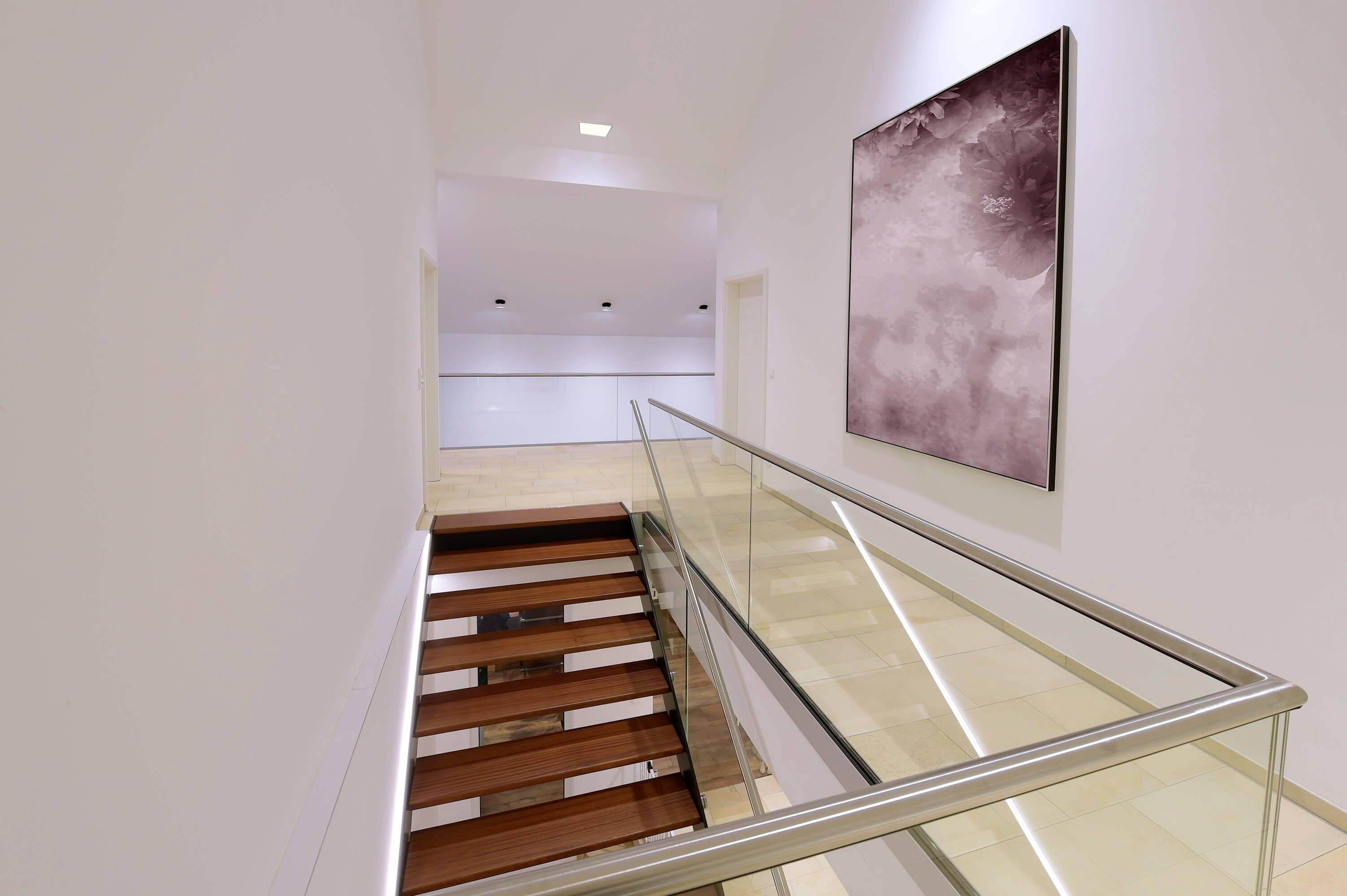 Couloir moderne avec une rampe en verre et un escalier en bois descendant. Une œuvre d'art abstraite en rose est suspendue au mur blanc.