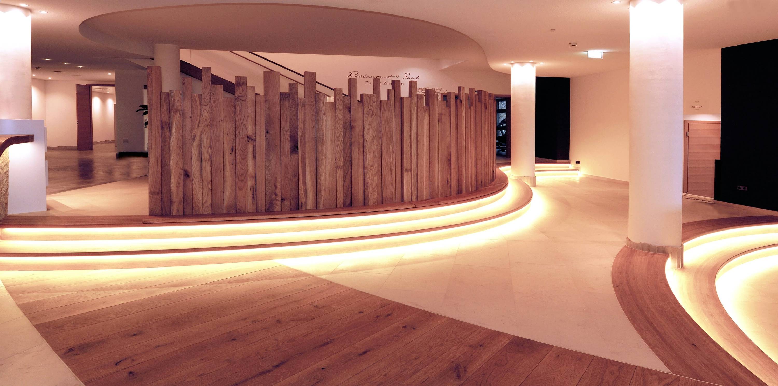 Un hall d'entrée moderne avec des sols en bois et en pierre éclairés. Un mur en bois semi-circulaire domine l'espace, baigné d'une lumière chaleureuse.