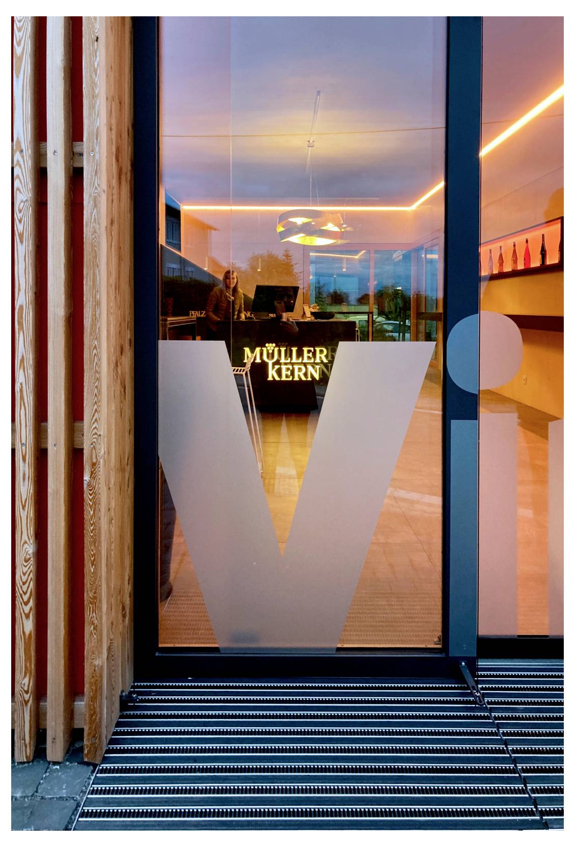 Entrée d'un bâtiment moderne avec une porte en verre ornée d'un grand « V », et « MÜLLER KERN » visible à l'intérieur