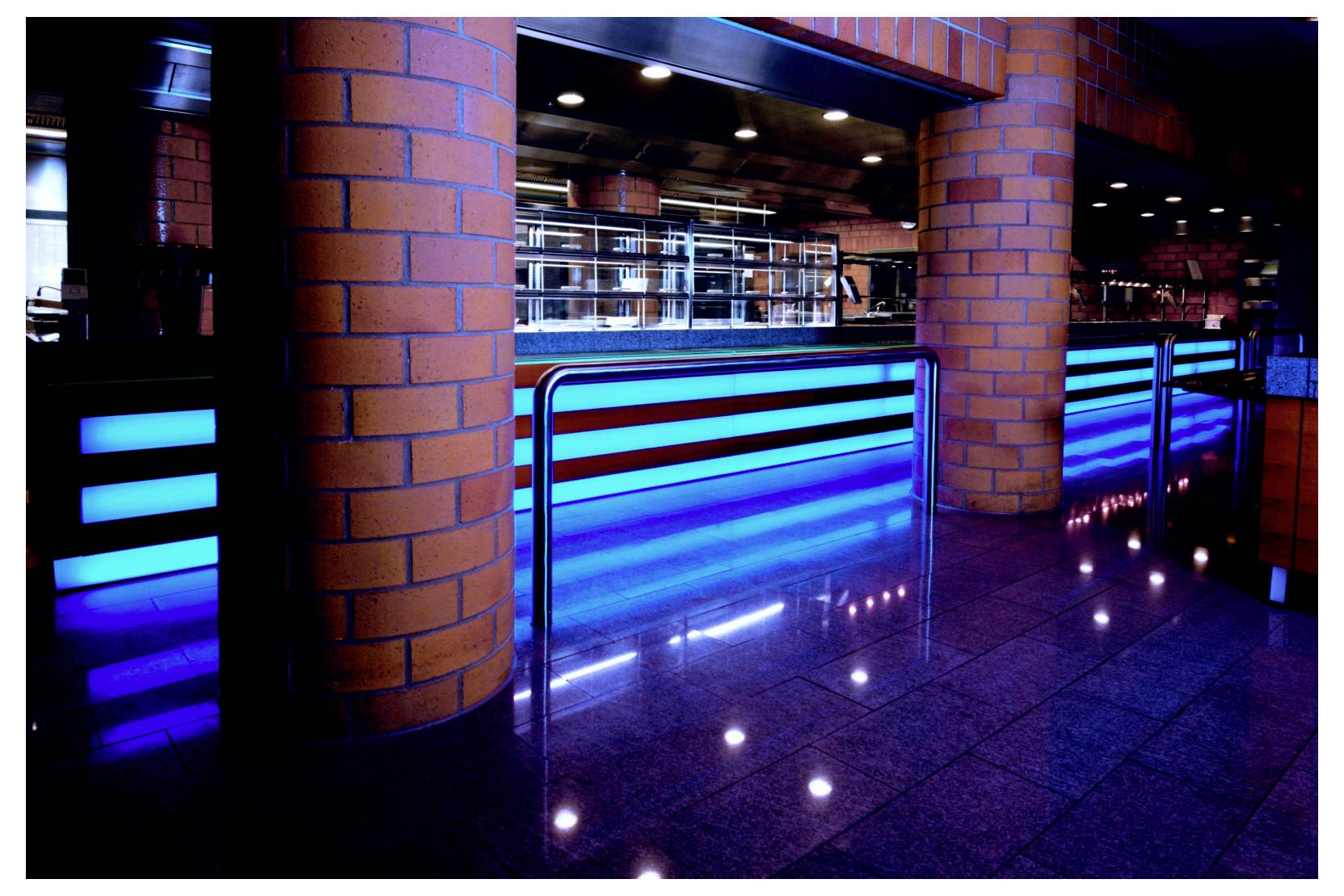 Intérieur d'une boîte de nuit ou d'un bar avec un comptoir éclairé en bleu, des colonnes en briques et un sol carrelé réfléchissant, vu depuis l'espace assises.