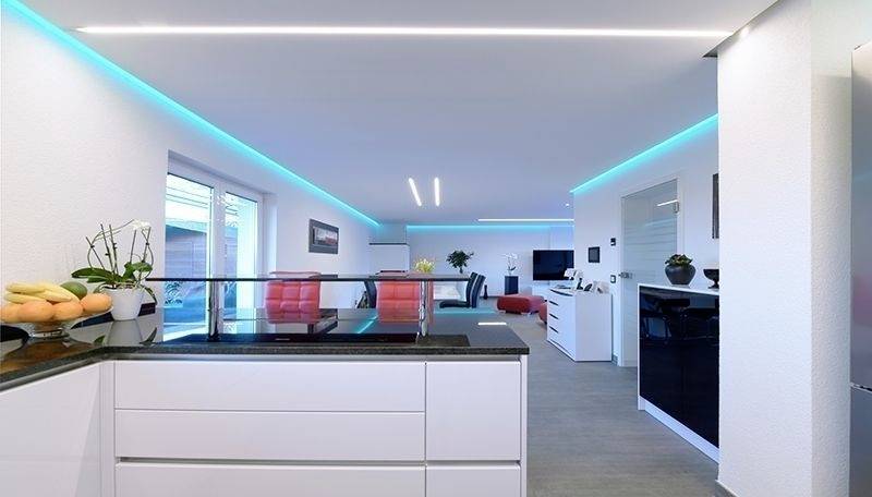Cuisine moderne avec un aménagement blanc, des plans de travail noirs et un éclairage LED bleu. Concept ouvert avec espace de vie en arrière-plan.
