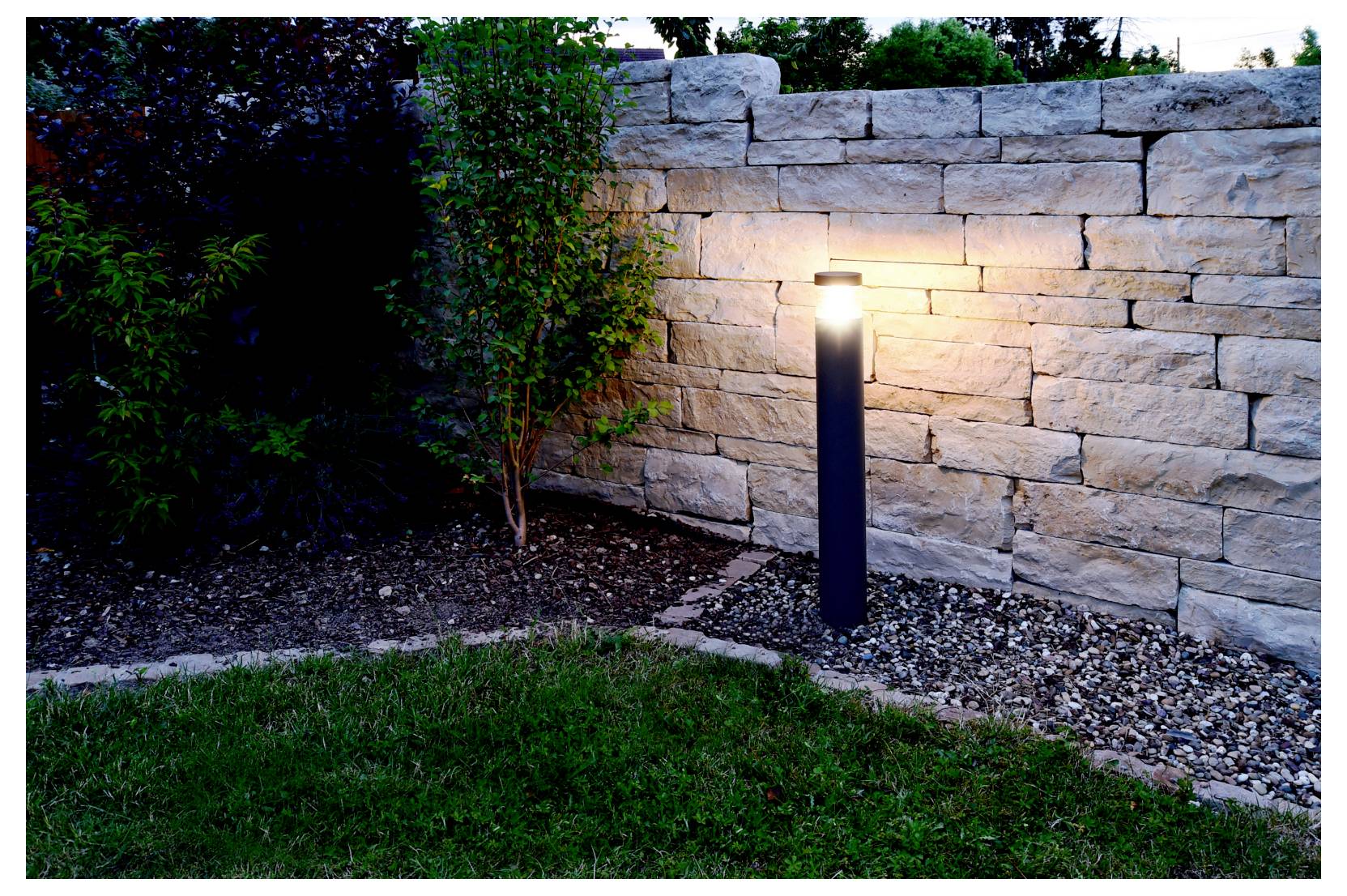 Poteau lumineux noir éclairé le long d'un mur en pierre, éclairant une plate-bande et la pelouse au crépuscule.