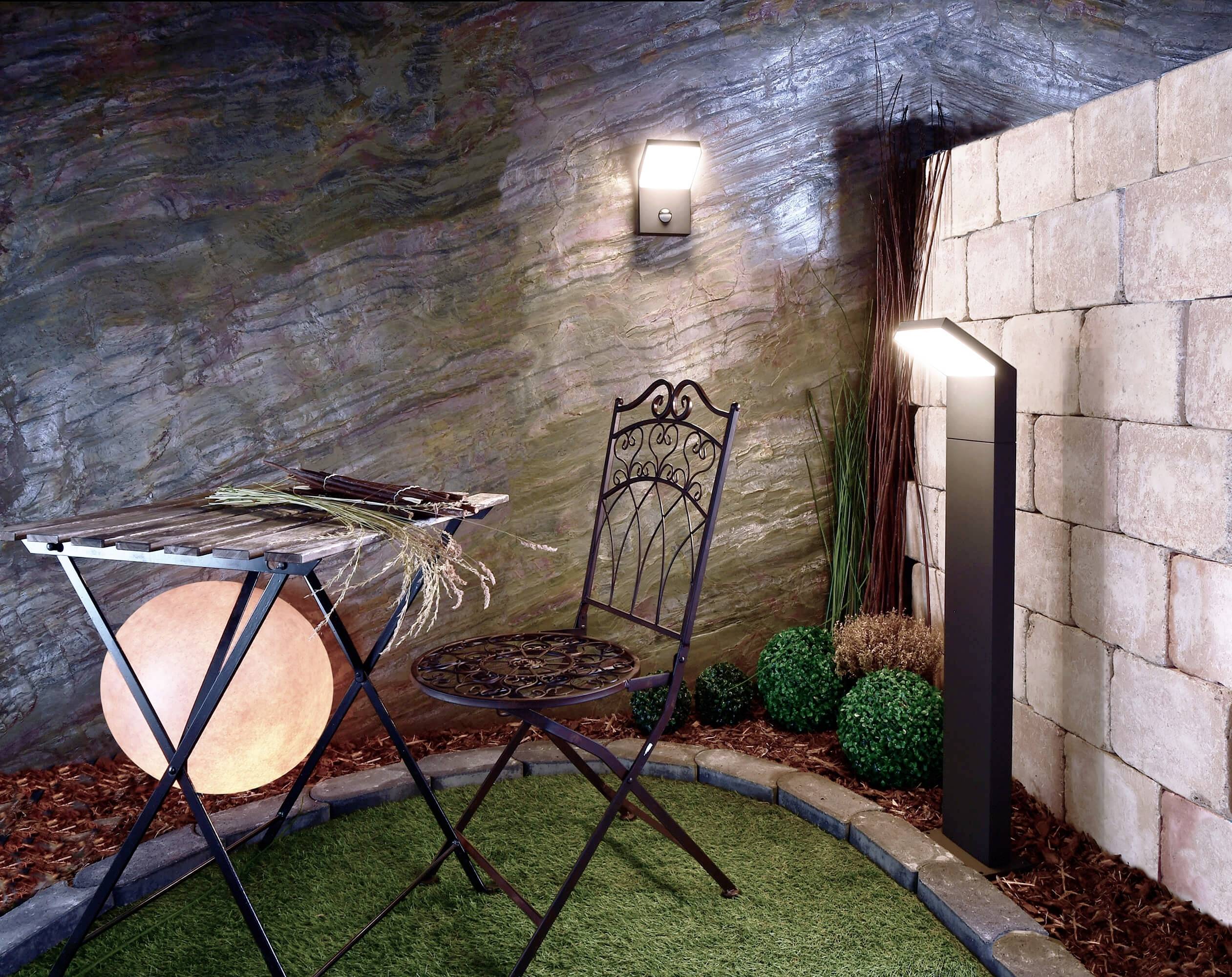 Un petit espace de jardin avec une table et une chaise en fer forgé sur un gazon synthétique. Une applique murale et un luminaire éclairent la scène.