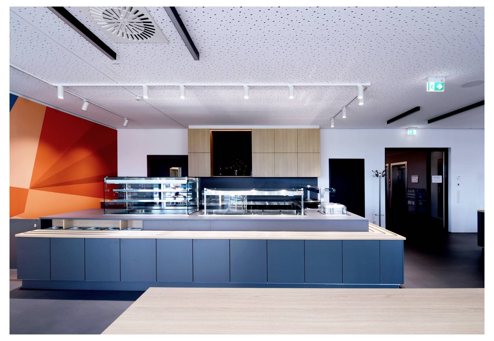 Cafétéria moderne ou comptoir de restauration avec vitrines en verre, meubles bleus, mur d'accent orange et éclairage sur rail au plafond.