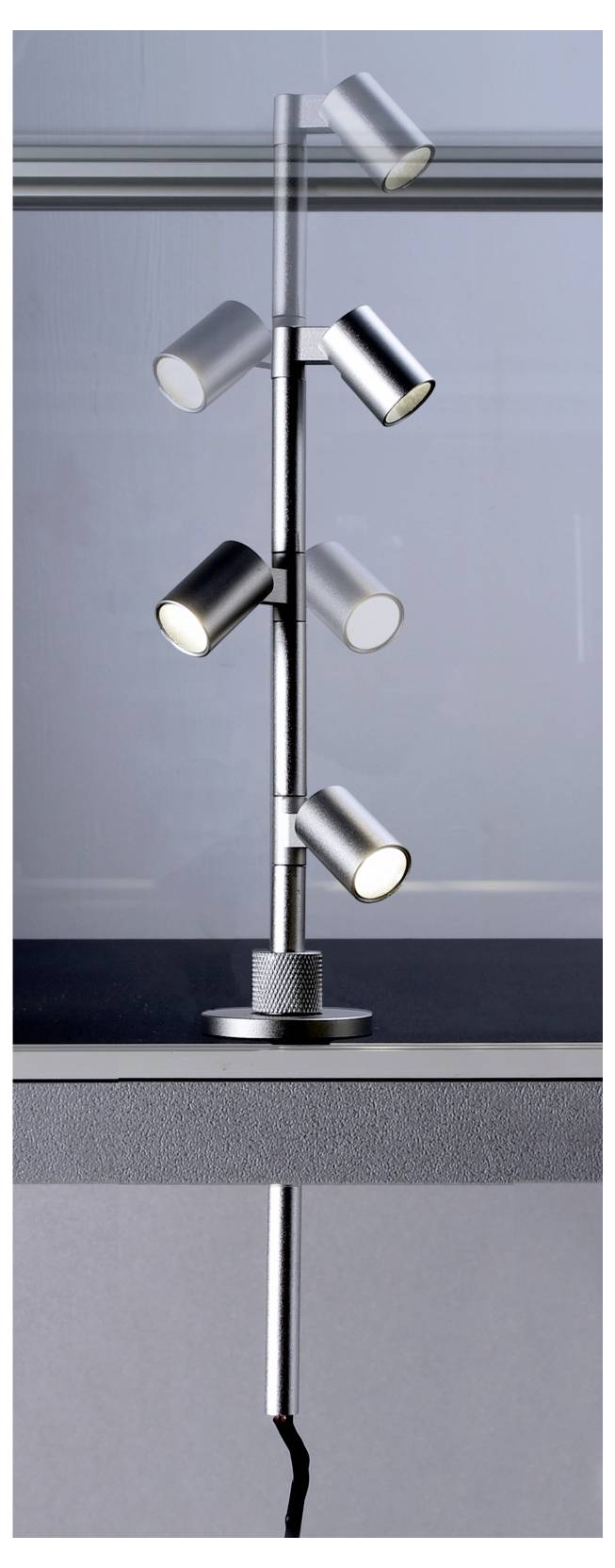 Lampadaire moderne à spots en chrome, doté de cinq têtes cylindriques orientables sur un fût fin et une base ronde, photographié devant un mur d'intérieur uni.
