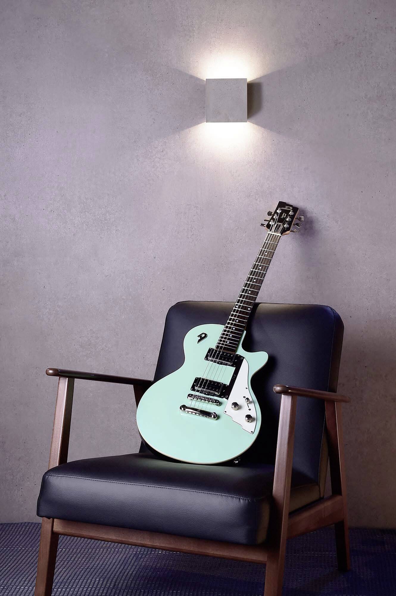 Une guitare électrique au corps turquoise est posée sur un fauteuil en cuir noir devant un mur gris avec une lampe murale lumineuse.