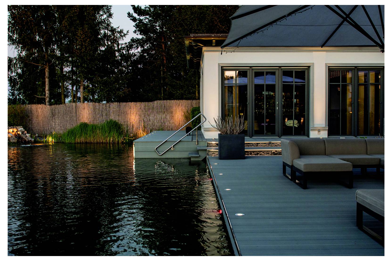 Terrasse moderne au bord d'un lac, près d'une maison au crépuscule, avec mobilier d'extérieur, marches éclairées menant à l'eau et vue sur une rive paisible.