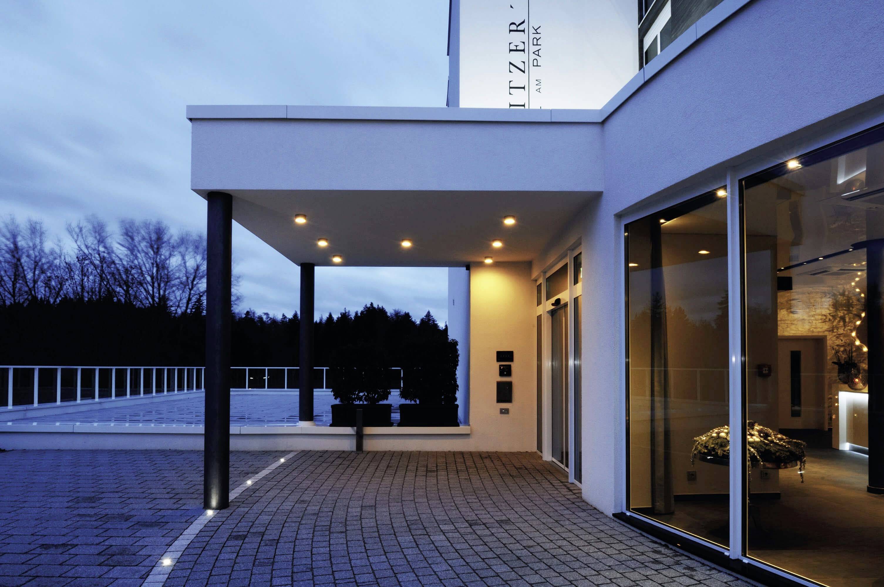 Un bâtiment moderne au crépuscule, illuminé d'une lumière chaleureuse. De grandes fenêtres révèlent un intérieur accueillant.