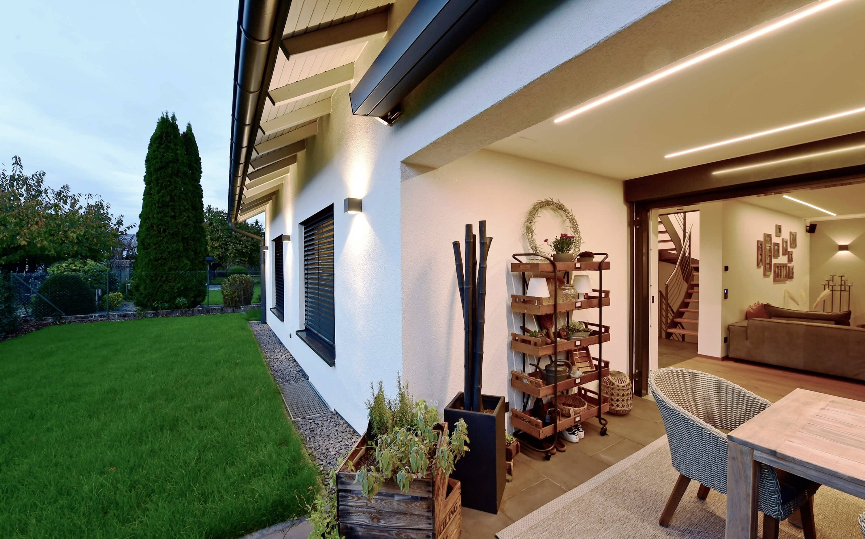 Une maison moderne avec une terrasse éclairée, une table et des chaises en bois. Le jardin est soigné, entouré d'arbres sous un ciel de soirée.