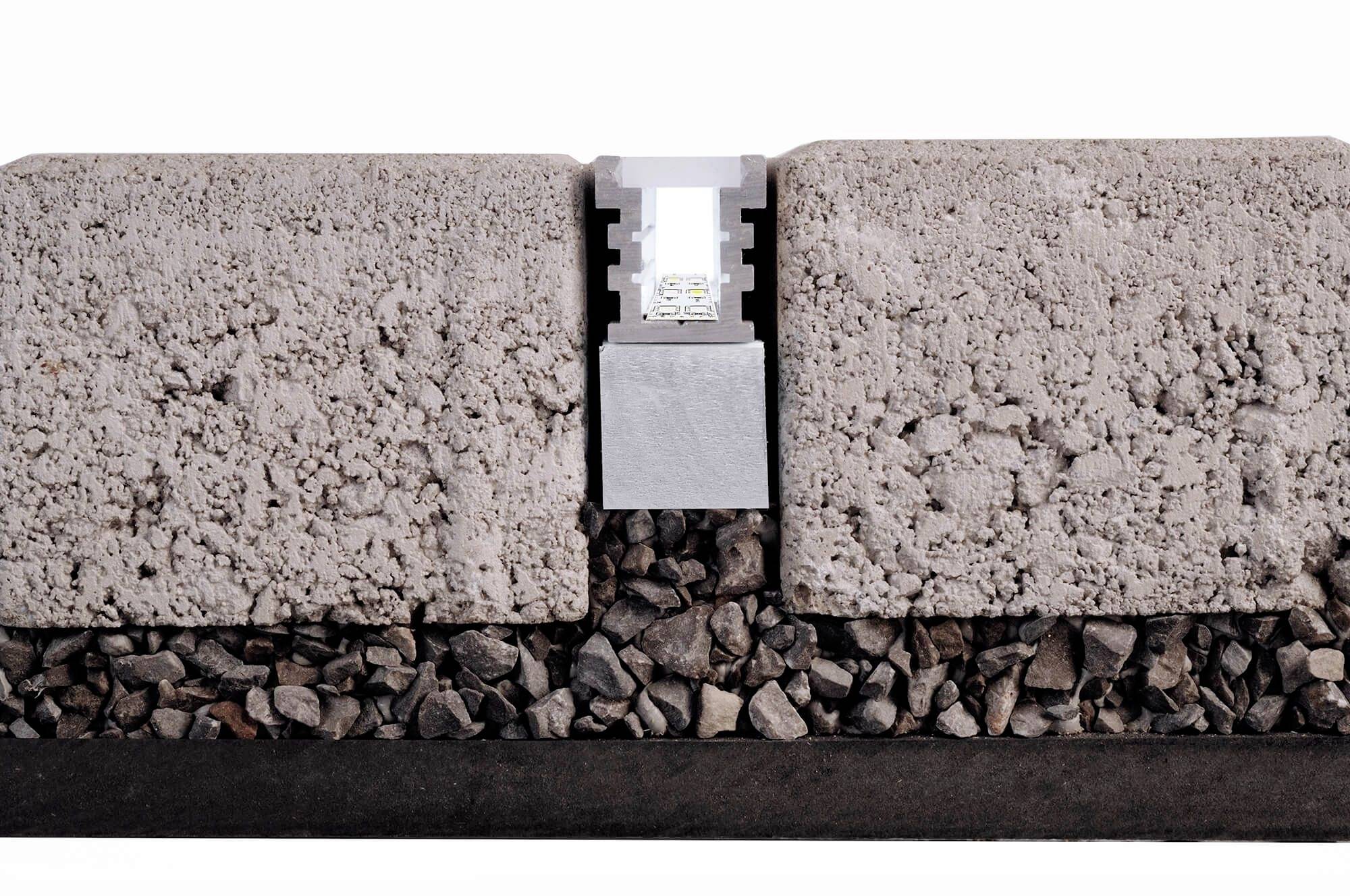 Des dalles de béton se rejoignent, séparées par une attache métallique. En dessous, se trouve une couche de petits cailloux.