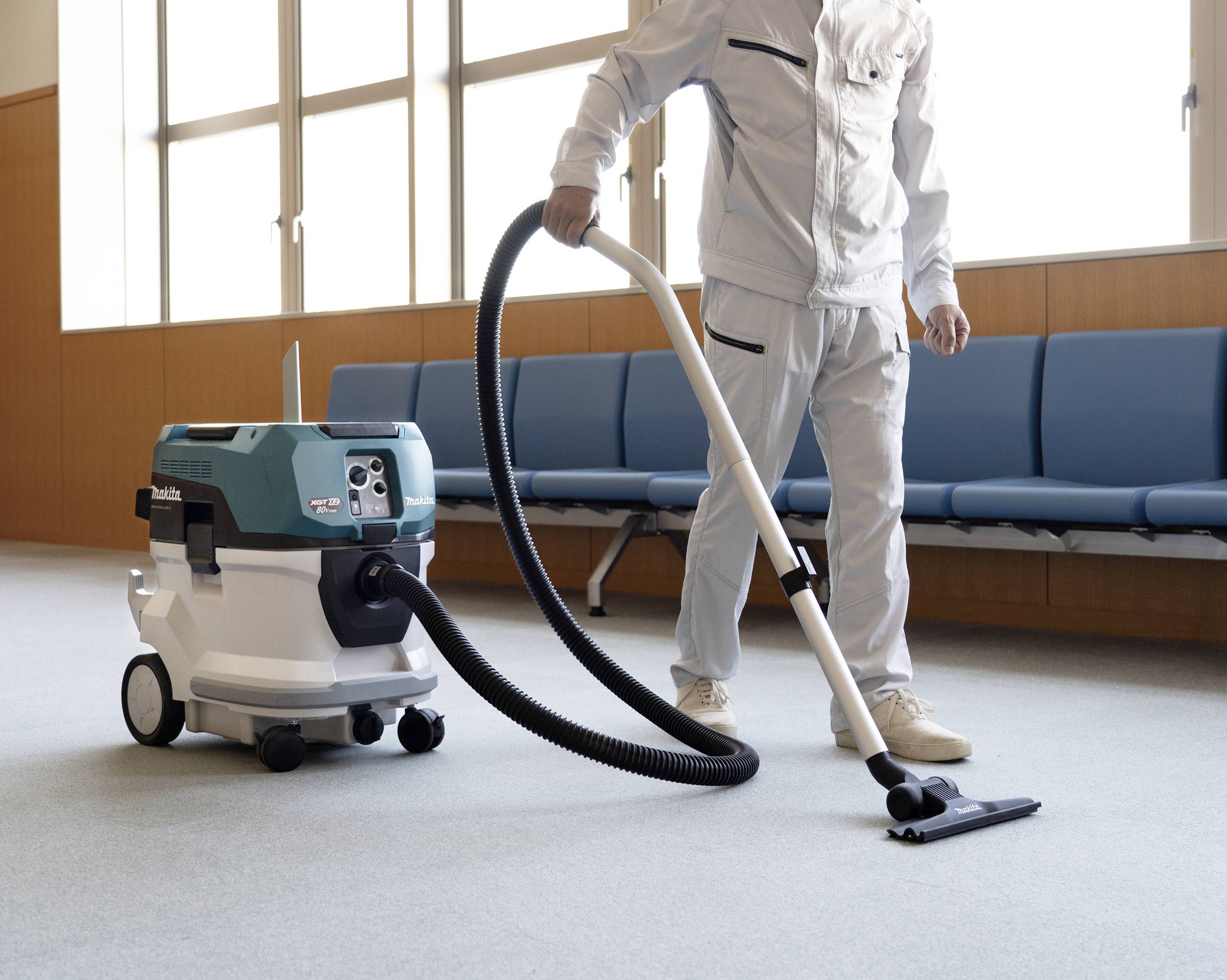 Une personne en tenue de travail utilise un aspirateur industriel sur un sol en moquette de bureau. Des chaises bleues sont visibles en arrière-plan.