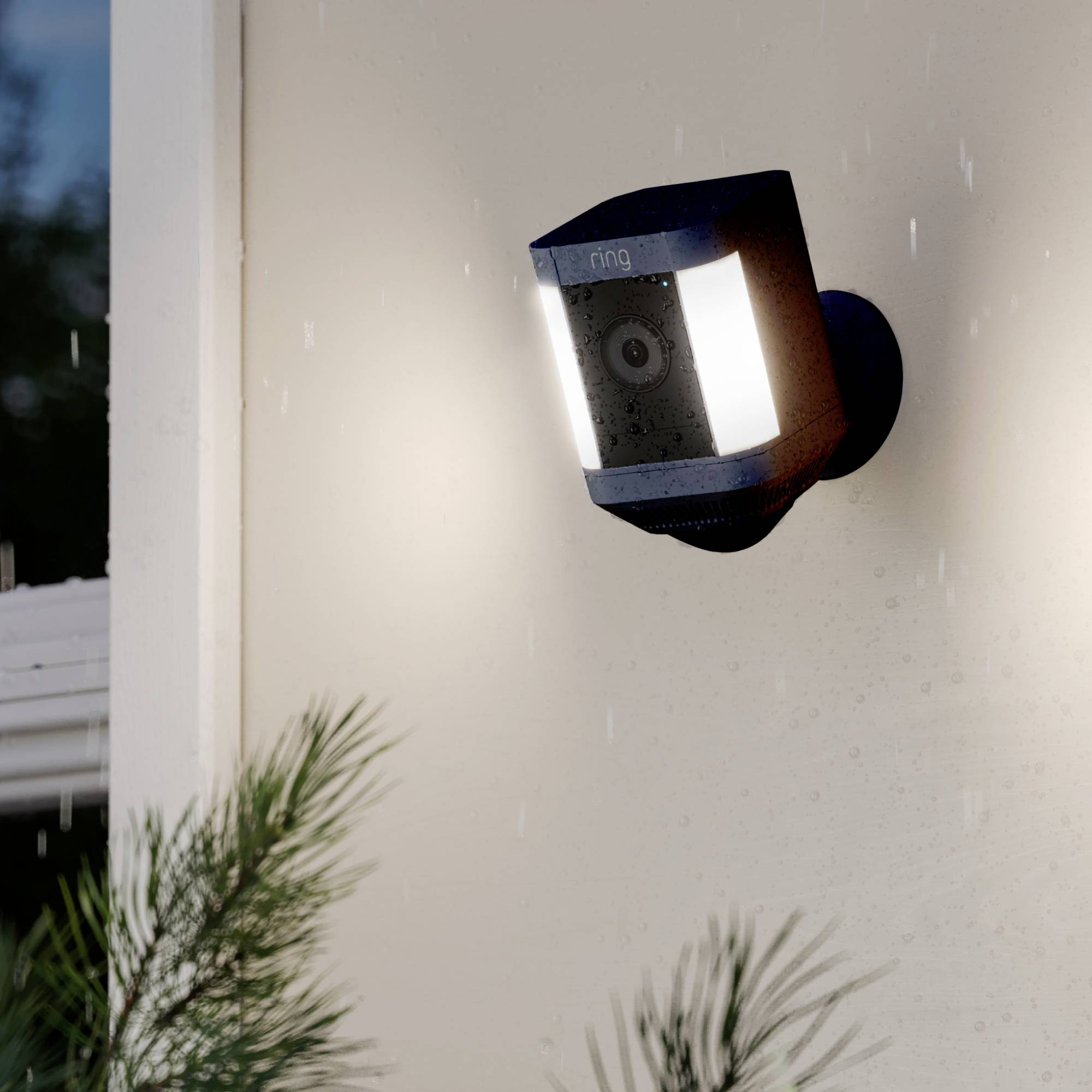 Une caméra de sécurité noire avec éclairage intégré est montée sur un mur de maison, entourée de pluie et de plantes au premier plan.