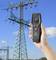 Une main tient un appareil de mesure numérique affichant des chiffres devant un fond comprenant un grand pylône électrique sous un ciel bleu.