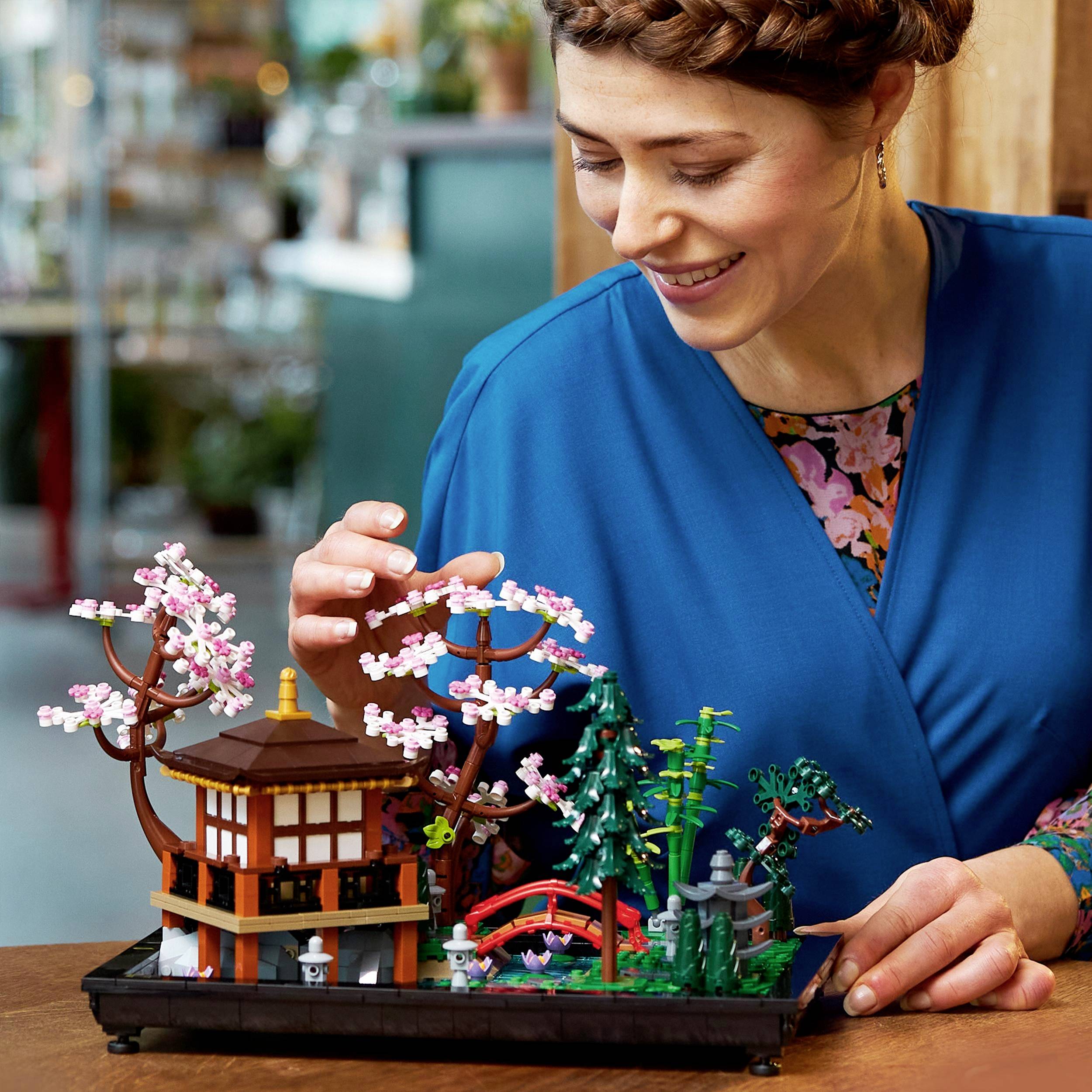 Une personne souriante observe une petite maquette d'un jardin japonais avec un cerisier en fleurs, un pont et un pavillon, le tout posé sur une table.