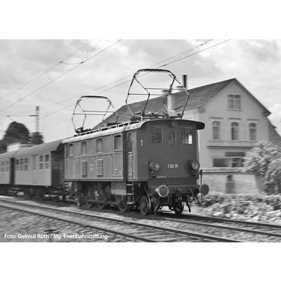 Une locomotive électrique historique tire un train sur une ligne ferroviaire rurale. Un bâtiment se trouve en arrière-plan.