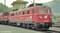 Locomotive électrique rouge de la série 1110 au toit gris, stationnée sur une voie devant une petite maison de garde-barrière. Des collines boisées sont visibles en arrière-plan.