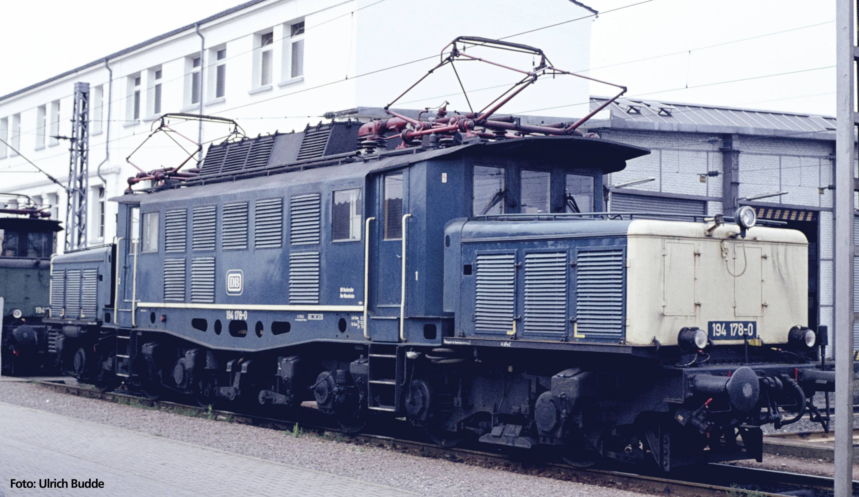 Une locomotive électrique bleue est stationnée sur une voie ferrée. Elle a des pantographes visibles en haut et un arrière-plan avec des bâtiments.