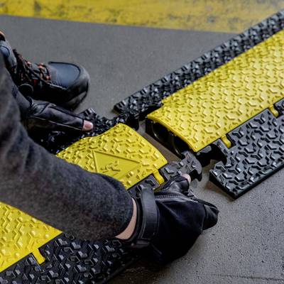 Une personne installe un tapis de protection au sol noir et jaune avec un motif antidérapant. Une tenue de sécurité est portée.