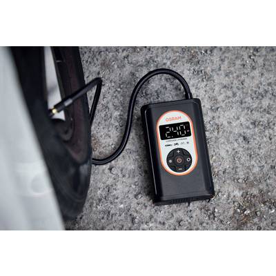 Un manomètre numérique de pression des pneus affiche '2,4 bar' et est connecté à une valve de pneu de voiture. L'appareil est posé sur du gravier.